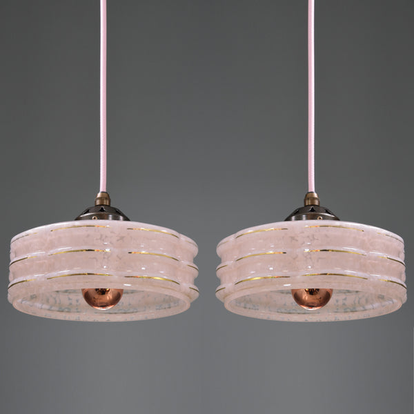 Two matching pendant lights with layered translucent pink shades, gold trim, and copper-toned bulbs hanging against a dark gray background.